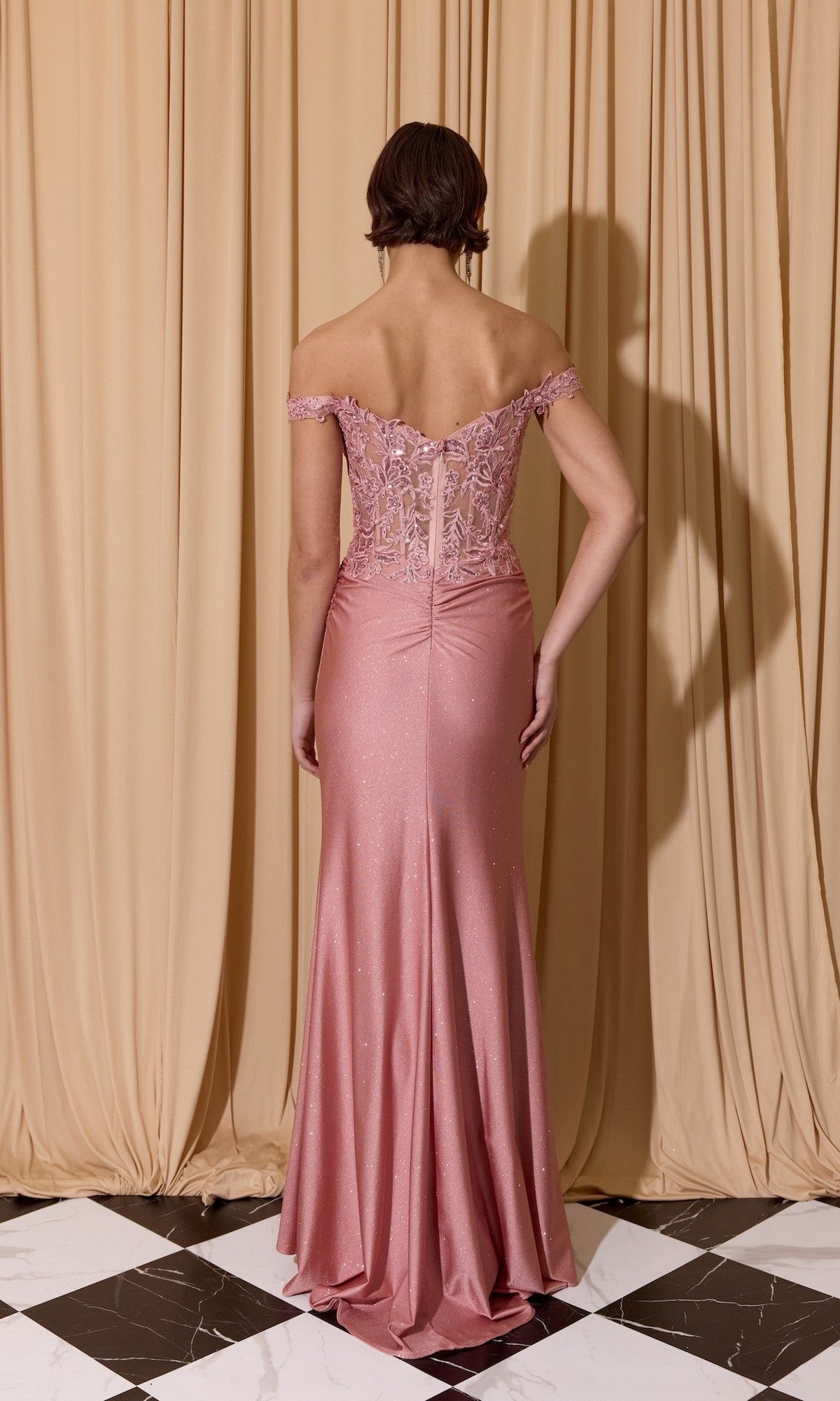 A woman poses with her back to the camera in the Eureka EK173 Long Prom Dress, which features a floral embroidered corset and flowing pink skirt, standing gracefully before beige curtains.